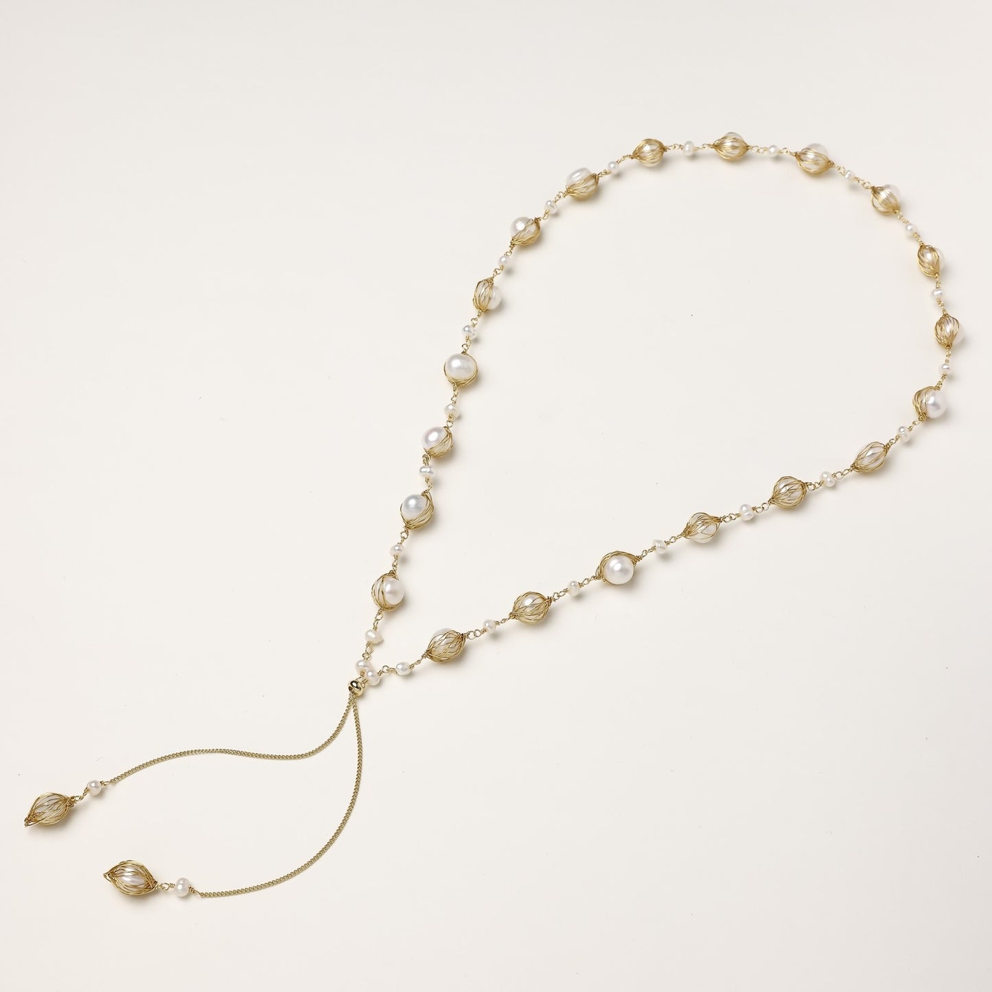 Long gold-plated lariat necklace with wire-wrapped freshwater pearls and chain tassel detail on cream background.