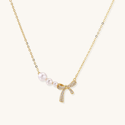 Delicate gold-plated necklace with three white pearls and sparkling bow pendant on cream background.