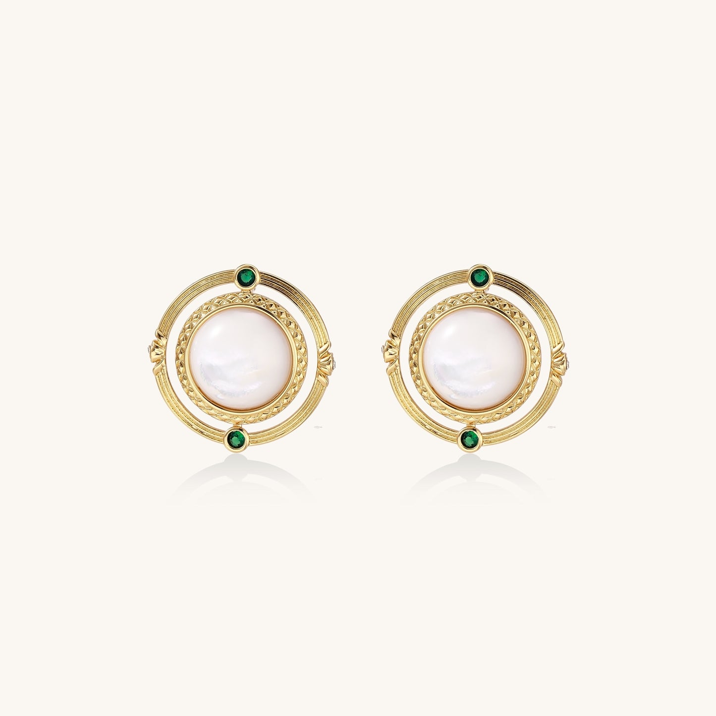 Close-up of gold-plated circular earrings with white mother-of-pearl centers and green zirconia accents on cream background.