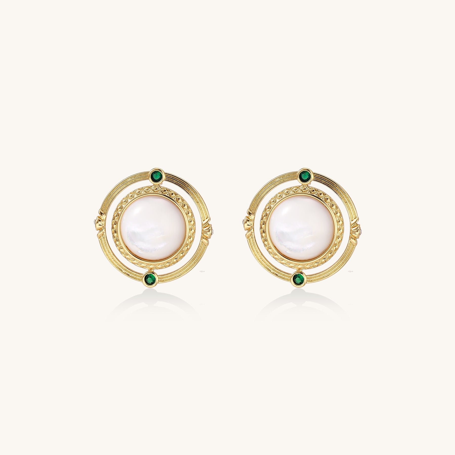 Close-up of gold-plated circular earrings with white mother-of-pearl centers and green zirconia accents on cream background.