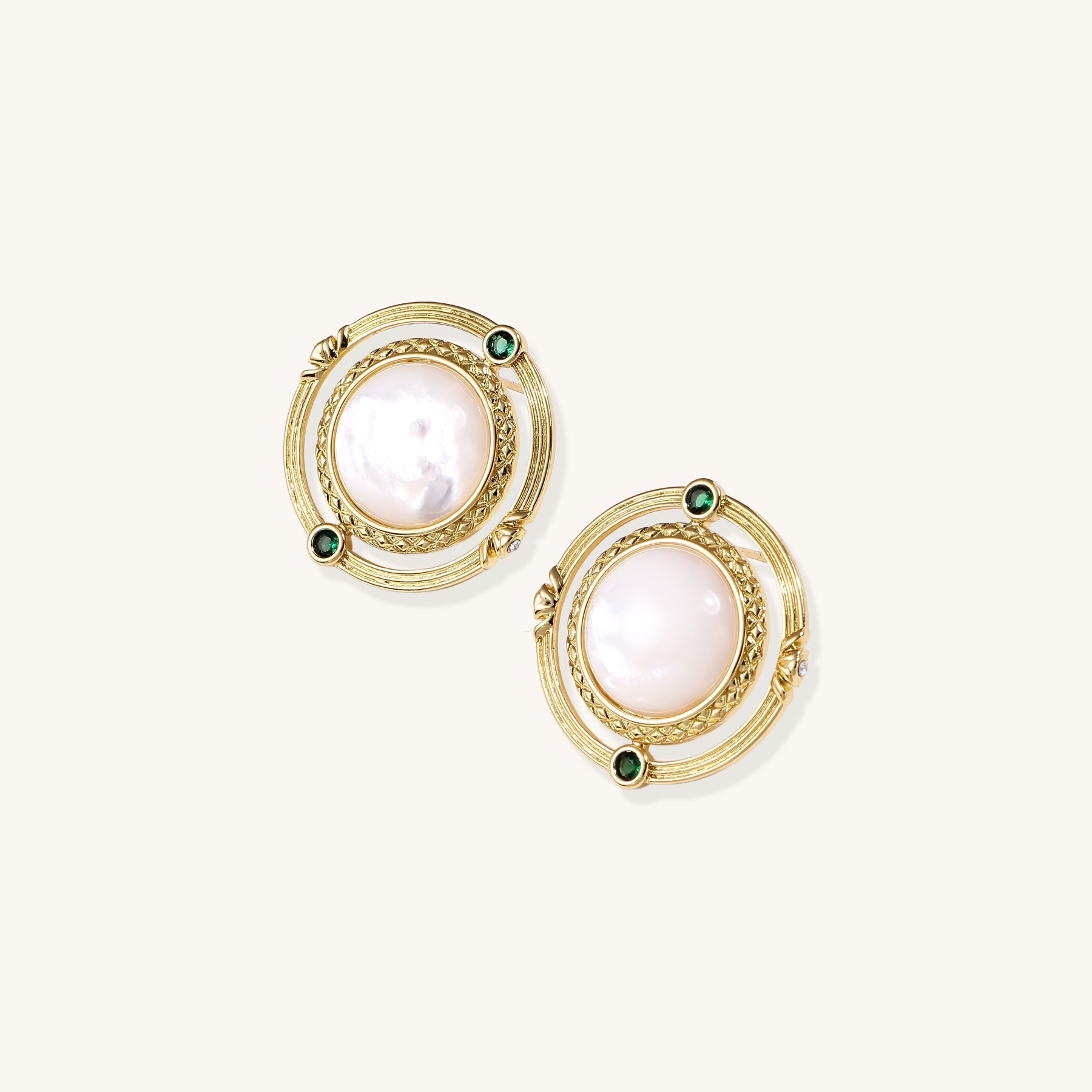 Close-up of gold-plated circular earrings with white mother-of-pearl centers and green zirconia accents on cream background.