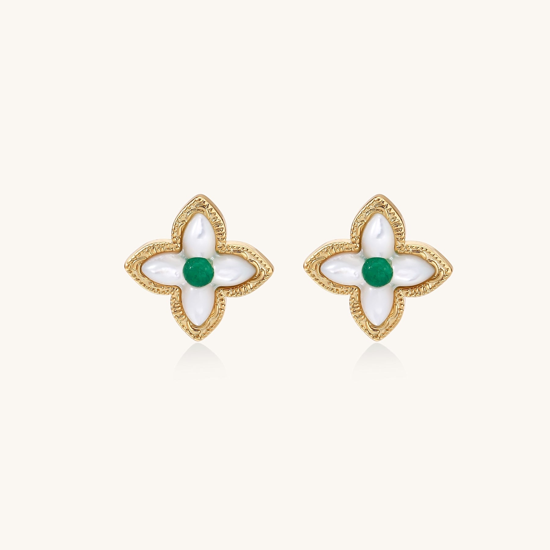 Dainty gold-plated quatrefoil stud earrings with white mother-of-pearl petals and green gemstone centers on beige background.