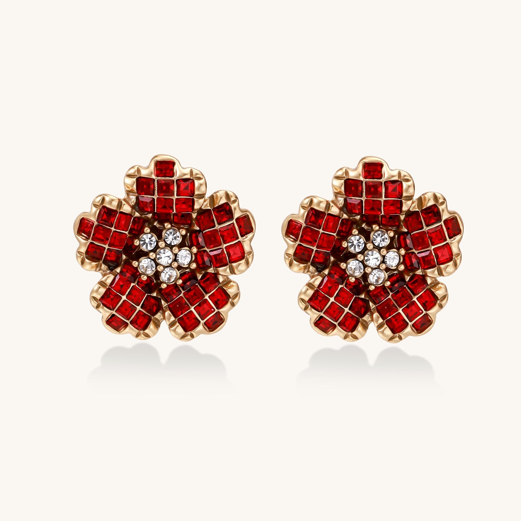 Gold-plated floral stud earrings with layered red crystal petals and clear zirconia centers on cream background.