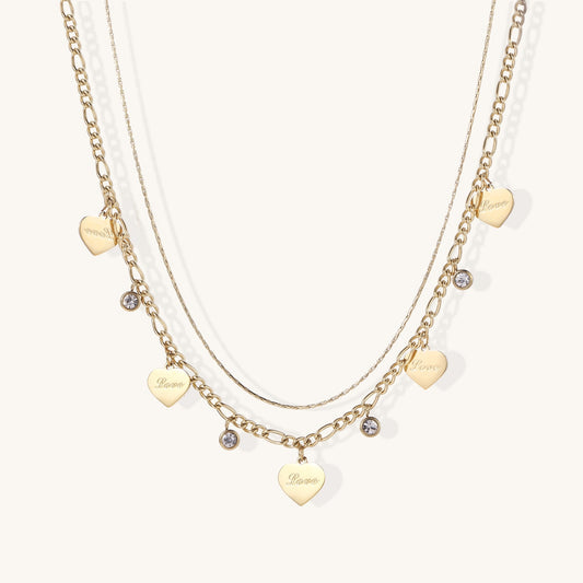 Two layered gold-plated necklaces with heart pendants engraved 'Love' and sparkling crystal charms on white background.