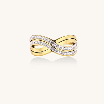 Gold-plated stainless steel ring with crossed infinity bands adorned with sparkling rhinestones against cream background.