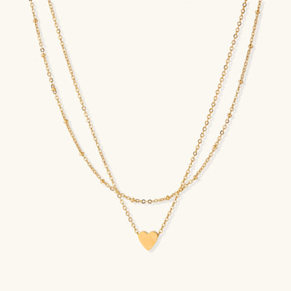 Two-layer gold-plated stainless steel necklace with small heart pendant displayed on beige background