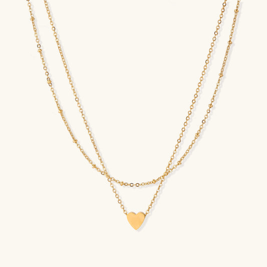 Two-layer gold-plated stainless steel necklace with small heart pendant displayed on beige background