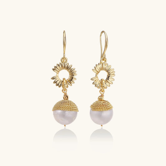 Gold sunburst charm earrings with baroque pearl drops on French hooks against cream background