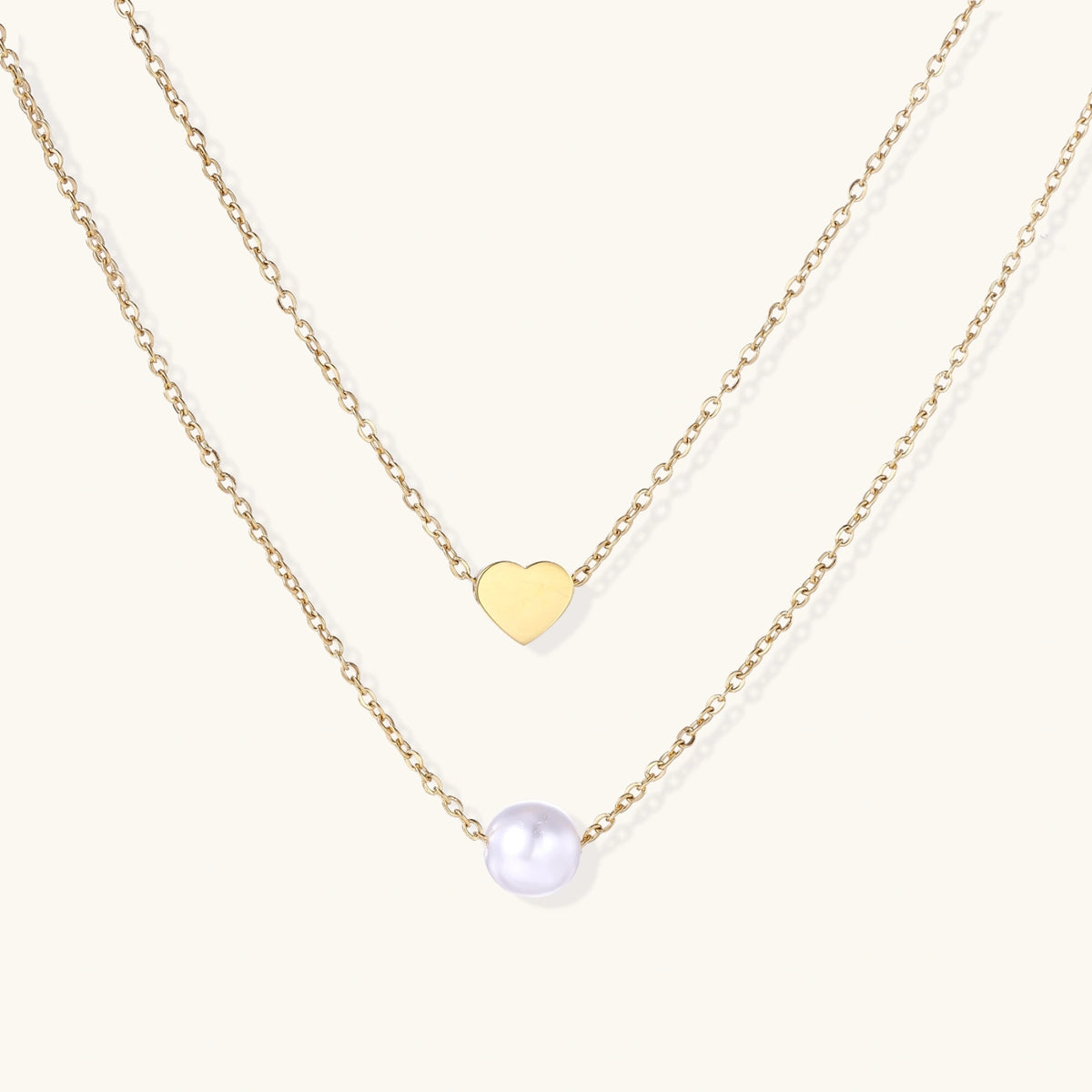 Two delicate gold-plated chains with heart charm and white pearl bead on cream background, minimal layered design.