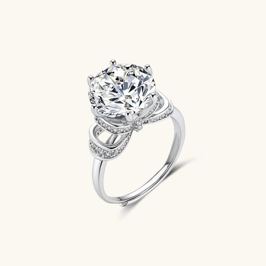 Sterling silver ring with 5ct round moissanite center stone and intricate bow design with pavé accents on white background.